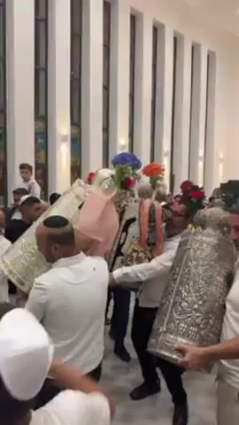 The video shows a religious celebration taking place indoors. Several men, some wearing yarmulkes, are carrying large, ornate silver objects adorned with flowers. They are surrounded by other people, including children, who appear to be dancing and clapping. The setting is a well-lit room with white walls and tall windows. A framed picture is visible on one wall. The atmosphere is lively and festive.