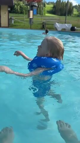 A young child wearing a blue inflatable flotation device around their neck is in a swimming pool. The child has blonde hair and is looking to the right, with their mouth open and a smile on their face. Their arms are raised and bent at the elbows, with their hands open. The water is clear blue, and the child's reflection is visible below them. In the background, there is another person in the pool, further away. Beyond the pool, there is a grassy hill with trees and a ski lift structure. The sky is partly cloudy. The camera appears to be positioned at water level, looking towards the child.