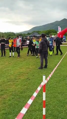 The video shows a group of people, likely participants in a race, lined up on a grassy field behind a red and white striped barrier. They appear to be preparing to start, with some looking at their phones or maps. Another group of people, potentially spectators, are standing behind another barrier in the background. There are some tents and an inflatable arch visible, possibly marking the starting or finishing point of the race. One person, wearing a green raincoat, stands near the barrier and appears to be addressing the participants. In the distance, there's a mountain range under a cloudy sky. The environment suggests an outdoor sporting event or competition taking place in a rural area.