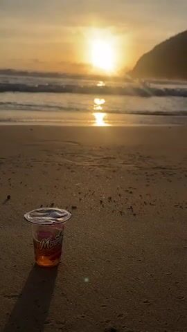 A plastic cup of jelly sits on a sandy beach at sunset. The sun is low on the horizon, casting a warm glow over the scene. Waves crash gently on the shore, and the silhouette of a hill can be seen in the distance. The cup has a clear plastic lid and appears to be orange or pink in color, with a label that reads "Mountain" in red lettering. The sand is wet and reflects the light from the setting sun. The overall mood is peaceful and serene.