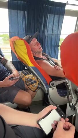 The video shows the interior of a bus. In the foreground, a person's hands are visible, holding a smartphone. In the middle ground, a man is asleep in a bus seat. He is wearing a black baseball cap, a black tank top, and has his arms crossed. His face appears red and flushed. The seat he is in has orange and yellow fabric on the headrest. A person on the left is wearing a dark blue t-shirt with white lettering, shorts, and has tattoos on their arm. They are holding a smartphone. Another seat with an orange and yellow cover is visible in the foreground to the right, facing away from the camera. The bus has blue curtains and some overhead compartments are visible, one with circular images. The overall environment appears to be a moving vehicle.