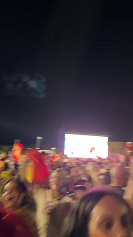 This is a vertical, blurry video of a crowd of people at night, appearing to be at an outdoor event. Many people are wearing red and yellow, and several are holding up Spanish flags. The atmosphere appears to be festive, with people waving flags and their arms in the air. There are bright lights visible in the background, suggesting a stage or stadium lighting. The overall impression is of a large gathering of fans, likely celebrating a sporting event or national holiday.