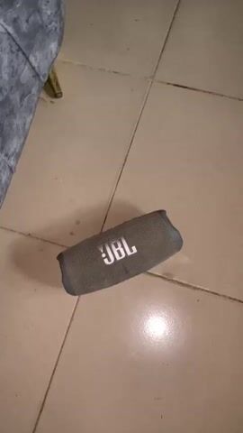 A gray JBL Bluetooth speaker sits on a tan tiled floor. A gray-covered chair rests partially in the frame to the left. A shadow is cast to the right of the speaker. Music is playing.