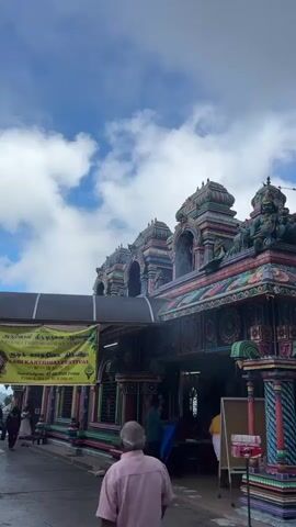 The video shows the exterior of a colorful Hindu temple under a cloudy blue sky. The temple is adorned with intricate carvings and sculptures in vibrant shades of pink, blue, green, yellow, and purple. Several people are visible, including a man in a pink shirt walking away from the camera, individuals standing near the entrance, and others seated on benches. A yellow banner with text is hanging on the left side of the temple. The architecture features multiple tiered roofs and decorative pillars. The overall scene suggests a place of worship with ongoing activity.