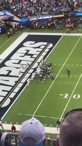 The video shows an American football game in progress, likely filmed from the stands. Two teams, one in white and the other in black, are facing off on a green field. The field features a large "CHARGERS" logo. The black team has the ball, and the players are engaged in a play. The crowd is visible in the background, filling the stadium. One person in the foreground is wearing a "TOMLINSON 21" jersey and cheering with arms raised. The overall environment suggests a lively and enthusiastic football game atmosphere.