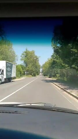 The video is recorded from inside a vehicle looking out through the windshield at a road lined with trees. The road is paved and has white lane markings. There are a few vehicles visible in the distance, including what appears to be a white truck or bus. There are also road signs visible, including what looks like a bus stop sign and a pedestrian crossing sign. The sky is clear and blue. The scene appears to be a rural or suburban road on a sunny day. The video is approximately 4 seconds long.