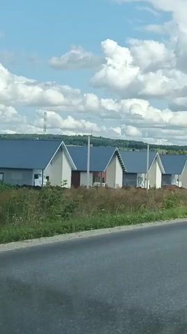The video shows a row of unfinished houses with gray roofs and light-colored walls. The houses appear to be in various stages of construction, with some having exposed brickwork and others with windows and doors. The houses are set against a backdrop of green grass, brown dirt, and a blue sky with white clouds. There are utility poles visible in the distance. The video is recorded from a moving vehicle, indicated by the slight blur and perspective shift. The overall impression is a suburban or rural setting undergoing development.
