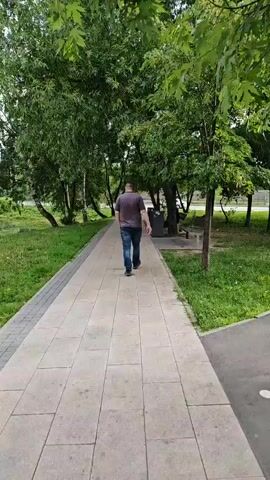 A person walks along a paved path in a park. The path is lined with grass and trees. The person is wearing a dark t-shirt and blue jeans. They are walking away from the camera. There is a trash can and a bench visible on the right side of the path. The trees are green and leafy, suggesting it is summer or spring. The sky is overcast.