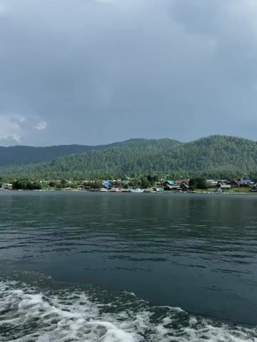 The video shows a wide shot of a large body of water with a village nestled along the shoreline in the distance. The water is dark and choppy, with white foam visible in the foreground, suggesting movement, likely from a boat. The village consists of various buildings, some with colorful roofs, scattered amongst trees. Behind the village, rolling green hills covered in dense forest rise up, disappearing into a hazy sky. The sky is mostly overcast with some lighter patches. The camera appears to be moving, offering a panoramic view of the landscape.