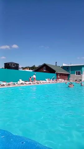 A wide shot captures a bright, sunny day at an outdoor swimming pool. The pool is filled with clear, turquoise water, and several people are enjoying a swim. The pool area is lined with lounge chairs, some occupied by people relaxing. In the background, a modern building with teal and white accents stands beside a smaller, rustic wooden structure. The sky is a clear, brilliant blue. The camera pans slowly to the right, revealing more of the pool and the covered seating area along its edge.