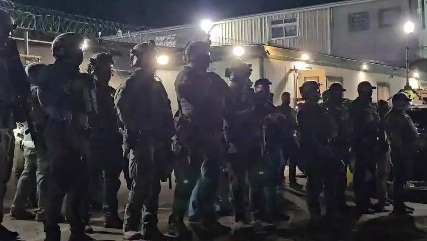 The video shows a large group of people, all wearing tactical gear and standing in formation in front of a building with barbed wire. They are silhouetted against the bright lights, and their faces are difficult to see. Some are wearing helmets, while others have caps. A few people in the back wear baseball caps. The building behind them has a window and appears to be a single-story structure. In the background, a police car is also visible. The people appear to be chanting or shouting something, but the audio is indistinct.