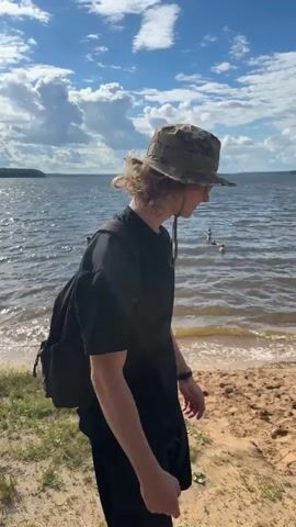 The video shows a person wearing a camouflage hat, a black shirt, and black shorts walking towards a lake. They are carrying a black backpack. The person is walking on a sandy path next to the water. The lake has waves and there are a few people swimming in the distance. The sky is blue with white clouds. The video is shot from behind the person.