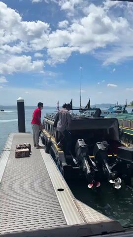 A man wearing a red shirt and gray pants stands on a metal dock next to a black and yellow boat. He appears to be assisting two people who are boarding the boat. Luggage is placed on the dock near the man. The boat has multiple outboard motors attached to its stern. Other boats are visible in the background, as is a blue sky with white clouds.