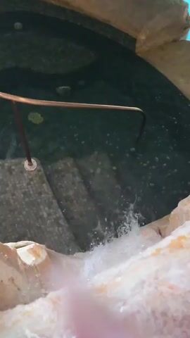 The video shows a small waterfall cascading into a pool. The pool has grey mosaic tile steps and a brown metal handrail. The surroundings of the pool are made of light-colored rock or stone. The water in the pool is a dark color and slightly murky. A few floating objects are visible on the surface of the water.
