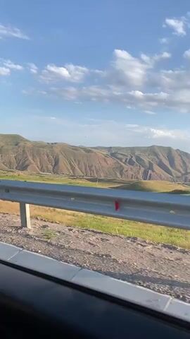 A side view from a car window captures a scenic landscape. In the foreground, a metal guardrail with red reflectors runs horizontally across the frame. Beyond the guardrail, dry grass and sparse vegetation are visible on a dusty roadside. The background features rolling, arid mountains with layered strata of brown and ochre. A bright blue sky with scattered white clouds completes the scene. The lighting suggests it is daytime, likely late afternoon due to the warm tones of the mountains and the angle of the light. There are no people or animals visible in the frame.