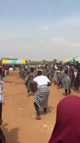 The video depicts a group of people performing a traditional dance in an outdoor setting. The dancers are dressed in black and white striped clothing, some with bare chests. They are moving in unison, performing coordinated steps and gestures. The scene is set on a dirt ground, with tents and a crowd of onlookers in the background. The video is shot from within the crowd, capturing the dancers from a low angle. The weather appears sunny. There is litter on the ground.