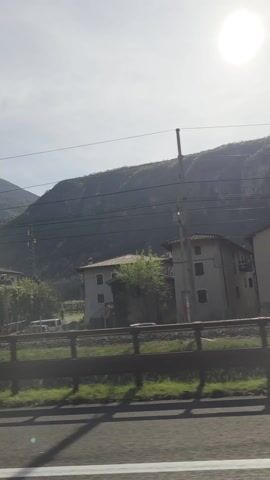 The view from inside a car shows a roadside landscape with mountains in the background. A railway track runs parallel to the road, with several buildings, likely houses, visible beyond it. Traffic signs are also present. The lighting is bright, with the sun causing a slight flare effect. A blue car passes in the same direction.