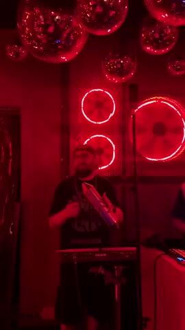 Two men are playing music in a room with red lighting. Disco balls hang from the ceiling. Behind them are lighted fan shapes. The man on the left plays a guiro and stands in front of a keyboard. The man on the right stands in front of a sound mixer and a computer screen.