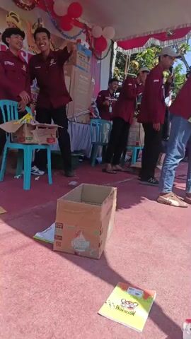 A group of young people, appearing to be teenagers or young adults, dressed in matching maroon shirts with a logo and black pants or jeans, are participating in an event on a stage. They are standing in a line, some holding microphones or speaking. The stage is covered with a red carpet and a white tent. There are balloons and decorations in the background. In the foreground, there are cardboard boxes and some papers. The overall atmosphere suggests a lively event, possibly a competition or performance. The subsequent clips show a street scene with people walking, some wearing traditional clothing. Indonesian flags are visible, suggesting a national celebration or holiday. There is a donation box labeled "MUSHOLLA AL-MIRADJI" and an inscription that appears to be in Arabic. The environment is outdoors, with buildings and trees visible. The sky is clear and sunny.