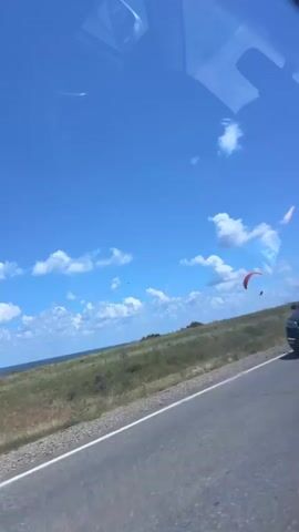 A paraglider with a red canopy and dark harness flies over a grassy field near the ocean under a blue sky with scattered white clouds. A white car is visible in the foreground on a road. The scene appears to be captured from within another vehicle.