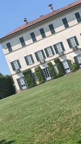 The media file captures a serene view of a grand villa and its meticulously maintained gardens under a clear blue sky. The villa, a prominent subject, is an elegant, multi-story structure with a cream-colored facade, accented by dark green shutters on its numerous windows. Ivy climbs artfully up the walls around some of the entrances, adding a touch of natural beauty to the architecture.

In front of the villa, a vast, verdant lawn stretches out, dotted with small white flowers, suggesting a pleasant, possibly late spring or summer day. The scene is peaceful and tranquil, with no people visible, allowing the focus to remain on the architecture and natural surroundings.

To the left of the villa, a tall, dense hedge forms a wall, with planters and a bench visible behind it, hinting at further structured garden elements. Further in the distance, the lawn continues towards another prominent feature: a long, elaborately shaped hedge, creating a formal garden design with archways. More planters line this hedge, and beyond it, a large, mature tree stands majestically, adding to the sense of lush greenery. In the far distance, a small fountain is visible, its water jet arcing into the air, adding a subtle element of movement and sound to the otherwise still imagery.

The time of day appears to be midday, indicated by the bright sunlight casting shadows on the lawn. The weather is clear and sunny, with a few wispy clouds in the expansive blue sky. The overall impression is one of quiet grandeur and timeless beauty, characteristic of a stately Italian villa estate. The location cues, such as the architectural style of the villa and the formal garden design, strongly suggest a European setting, consistent with Varese, Italy. There is no visible text in the media file.