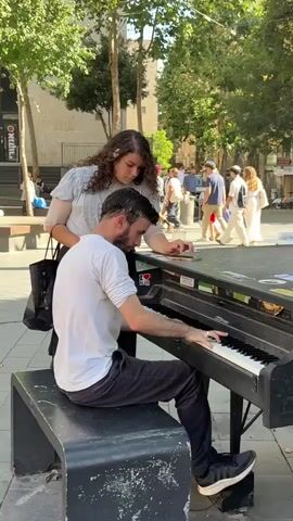 A man with a beard and a white t-shirt is playing a piano outdoors. He is sitting on a black bench in front of the piano. A woman with curly brown hair and a light-colored top is standing behind him, looking at the piano keys and holding a phone. She appears to be touching the piano. There are people in the background, some walking and some sitting. There are trees with green leaves and buildings in the background. The ground is paved. The lighting suggests it is daytime.