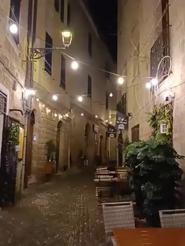 The video begins with a view of a narrow, cobblestone alleyway at night. String lights are hung across the alley, illuminating tables and chairs set up outside what appear to be restaurants or cafes. The buildings on either side are made of stone, with windows and signs visible. There are plants in planters along the sides of the alley. As the video progresses, the camera pans along the alley, showing more of the outdoor seating and the buildings. Several people are seen walking through the alley and sitting at the tables. In the latter part of the video, a sign with the word "Napoli" is visible on an awning. The overall atmosphere is that of a lively, European-style street at night.