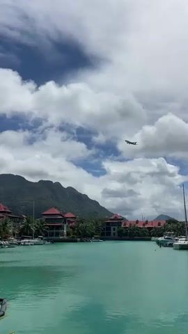 A small airplane flies across the sky over a body of turquoise water. Several boats are docked in the water, and buildings with red roofs line the shore. Lush green mountains are visible in the background under a cloudy sky.