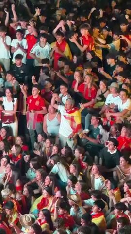 A high-angle, vertical shot captures a dense crowd of people, mostly young adults and adolescents, packed together. The majority of the individuals are wearing clothing in shades of red and yellow, some with Spanish flags draped over their shoulders or bodies, suggesting a patriotic or sporting event. The crowd is looking upwards and to the left, with many people cheering, raising their hands, and clapping. The lighting appears to be artificial and somewhat dim, creating a contrast with the bright colors of the clothing. There are glimpses of trees in the background, indicating an outdoor setting, possibly a public square or stadium seating area at night. The overall atmosphere is one of excitement and collective engagement.