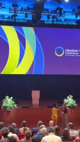 The video shows a conference room with a large screen displaying "Ukraine Recovery Conference 11-12 July 2022 Lugano, Italy and Ukraine." In front of the screen is a stage with a white podium, flanked by floral arrangements. The audience is seated in rows. Two women, one in a yellow suit and the other in beige pants, are on the stage talking. The woman in the yellow suit moves toward the left of the screen, and a woman in a black dress moves into a similar position after her. The audience appears to be watching the stage. The lighting is bright, and the overall atmosphere seems formal.