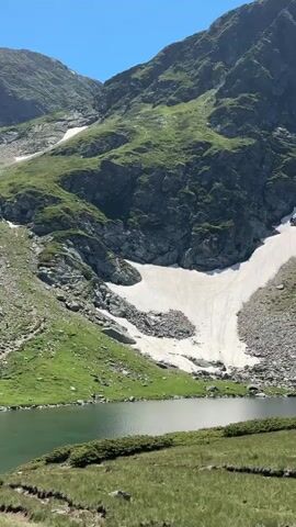 A vertical video shows a mountainous landscape with a lake in the foreground. The mountains are covered in green grass and patches of snow, with rocky outcrops. A trail winds along the side of the lake and up the mountainside, with a line of people walking on it. The lake water is green and reflects the sky. The sky is blue and clear. The video pans upwards from the lake towards the mountains, revealing more of the hiking trail and the snow patches.