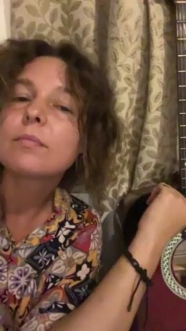 A woman with curly brown hair is featured in a close-up shot, looking directly at the camera. She is wearing a colorful, patterned shirt. Her right arm is visible, adorned with a black braided bracelet and a ring on her right ring finger. She is holding a maroon-colored acoustic guitar, which is positioned to the right of her in the frame. Her right hand is resting on the guitar's strings near the soundhole, and she is gently plucking individual strings throughout the video. The background consists of a light-colored curtain with a subtle dark leaf pattern. The woman maintains a calm expression, offering a slight smile towards the end of the video. The scene appears to be set indoors.