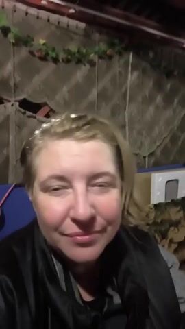 A blonde woman wearing a pearl headband and a black jacket looks into the camera with a slight smile. She is in an indoor setting with a patterned curtain behind her and some objects to her right. The lighting is dim.