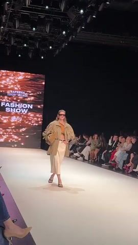 A fashion show runway is depicted with a model walking towards the camera. The model is wearing a tan jacket, an orange top, and a light-colored skirt, with sunglasses and a purse. In the background, an audience is seated on the right, and a large screen on the left displays "FASHION SHOW" in white text over a sparkling, amber background. The lighting suggests an indoor event space.