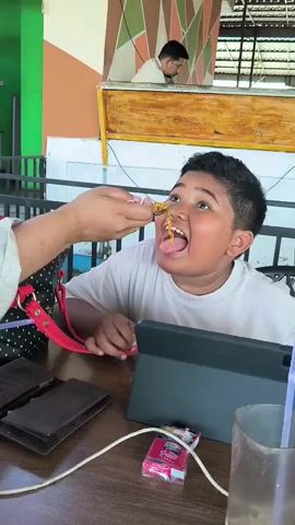 Here's a concise description of the video:

A young boy is sitting at a table, occupied with a tablet. Someone is feeding him noodles with a spoon. There is a wallet, a pack of cigarettes, and a glass with a straw on the table. Another man is visible in the background, inside a building. The overall setting appears to be an outdoor restaurant or patio.
