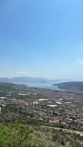 The video is a panoramic view of a coastal city with a bay and surrounding hills. The sky is clear blue with a few wispy clouds. In the foreground, there are dry, grassy hills with scattered green bushes and trees. The city is spread out below, with red-tiled roofs and white walls. A bay or harbor is visible in the middle ground, with several boats and yachts docked. Further in the distance, there are more hills and islands. A large white ship is visible sailing in the bay. The overall impression is one of a sunny, warm day in a Mediterranean coastal town.