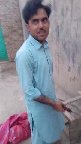 The video begins with a medium shot of a man standing in an outdoor courtyard. He is wearing a light blue shalwar kameez and has a mustache. He is looking at the camera and smiling. In the background, there is a brick wall and a green wooden door. The man is holding a cooking utensil in his right hand. The camera then pans down to show a cooking scene. A pot is placed over an open fire, with flames visible. The pot contains what appears to be food being cooked. There is smoke rising from the pot. A wooden stool is placed next to the fire. The ground is dirt and cracked. There are also some pieces of wood and debris on the ground. A red cloth is partially visible in the lower left corner of the frame. The video appears to be a casual recording of someone cooking outdoors.
