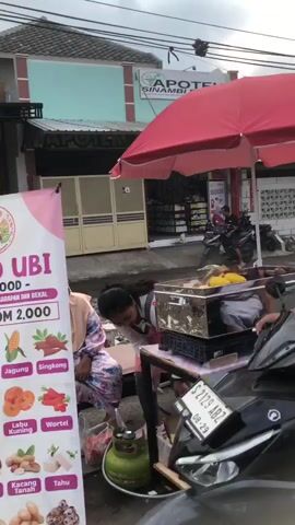The video opens with a close-up shot of the handlebars and front of a black motorcycle. A person, seen from behind and wearing a black t-shirt, is holding the handlebars. The camera pans slightly to the left, revealing a street scene. In the foreground, a food cart is visible, covered by a large red umbrella. Several food items, appearing to be various types of snacks, are displayed on the cart. To the left of the food cart, a woman wearing a light-colored hijab and a patterned dress is sitting on a low wall or curb. She appears to be interacting with the food cart. Behind the food cart and the woman, a white SUV is parked on the side of the street. Further in the background, a building with a closed doorway is visible, and another person, dressed in dark clothing, can be seen standing near the doorway. The overall environment suggests an outdoor street setting, possibly a marketplace or a roadside stall.