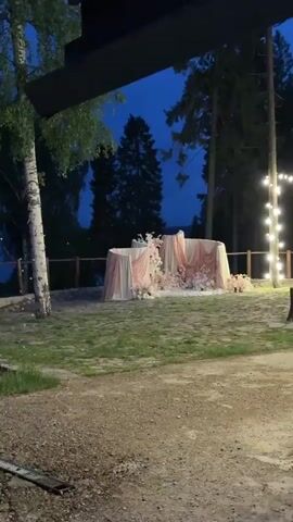 The camera captures a dark outdoor scene at dusk, showing a wedding arch decorated with pink and white flowers and flowing fabric. The arch is composed of several rounded panels draped with light pink fabric and adorned with clusters of pink and white flowers at the base and sides. The ground in front of the arch is a mix of dirt and sparse grass. In the background, silhouetted trees stand against a deep blue sky. No people are visible. The lighting is dim, indicating either early morning or late evening.