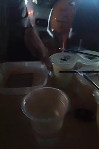 A young person wearing a dark shirt sits outside at night, preparing drinks. There are several small plastic cups with black lids and straws on the table. The cups have a logo featuring a stylized man with glasses. The person is filling a large white plastic container with something from another container.  Traffic and lights are visible in the background.