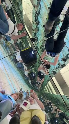 The vertical video captures multiple people moving within an immersive mirrored room, creating an illusion of infinite reflections on all surfaces – floor, walls, and ceiling. The primary focus is on a woman in the foreground wearing yellow pants, a white t-shirt, and a white baseball cap, who is holding a smartphone and appears to be recording the experience. She is smiling and looking towards the camera. Another woman in dark blue jeans and a white and blue striped top is seen walking through the space. Various other individuals are visible in the reflections, some holding phones themselves. A small black device is seen on the mirrored floor. The reflections multiply the figures and the room, creating a disorienting yet captivating visual effect. The scene appears brightly lit, suggesting an indoor exhibit or art installation in Haría, Spain.