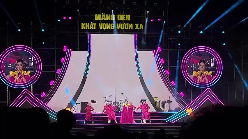 This image captures a vibrant night concert or festival performance on a large, elaborately lit stage. The setting is Măng Đen, Vietnam, as indicated by the prominent text "MĂNG ĐEN KHÁT VỌNG VƯƠN XA" displayed on the main LED screen, which translates to "Măng Đen - Aspiration to Reach Far."

On stage, a group of performers, primarily women, are engaged in singing and dancing. They are all dressed in striking, bright pink traditional Vietnamese-inspired outfits, some featuring flowing capes or scarves, and a few wear conical hats (nón lá). In the background, a band with a drum set and other instruments is partially visible, providing live music.

The stage itself is a focal point, featuring a large central LED screen flanked by two smaller, circular LED screens on either side. These side screens display close-up live feeds of a female vocalist, also in a pink outfit. The stage is framed by a dynamic arrangement of bright neon-like LED strips in pink, blue, and white, forming geometric patterns that create a modern and energetic aesthetic. Overhead and around the stage, numerous spotlights illuminate the performers and the intricate stage design.

In the foreground, a silhouetted audience watches the performance, adding to the atmosphere of a large public event. The overall impression is one of a lively, culturally rich celebration taking place under the night sky.