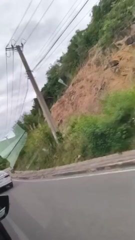 The video begins with a view from inside a vehicle, looking out the side window at a hilly, vegetated landscape. A white car is visible driving on a road next to the vehicle. Power lines run overhead, supported by a utility pole. The scene shifts to the interior of the vehicle, showing the dashboard, steering wheel, and a driver's hand. The windshield displays a road ahead with other vehicles, surrounded by green, tree-covered hills. A navigation screen on the dashboard shows a nature scene. A traffic sign with a red circle and horizontal bar is visible on the right side of the road.