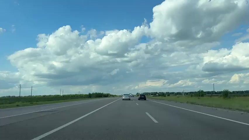 This is a first-person perspective video of driving on a highway under a bright blue sky with large, fluffy white clouds. The road is asphalt and has clear white lane markings. There are several cars visible on the highway, moving in the same direction. On either side of the highway, there are fields and trees. 

In the distance, there are power lines. The weather appears to be pleasant, possibly a warm day. The time of day seems to be late morning or early afternoon, given the bright sunlight and the angle of the shadows.

There are no people visible in the frame, but the presence of cars suggests human activity. The scene is typical of travel on a highway in a rural or semi-rural area. There is no text visible in the video.