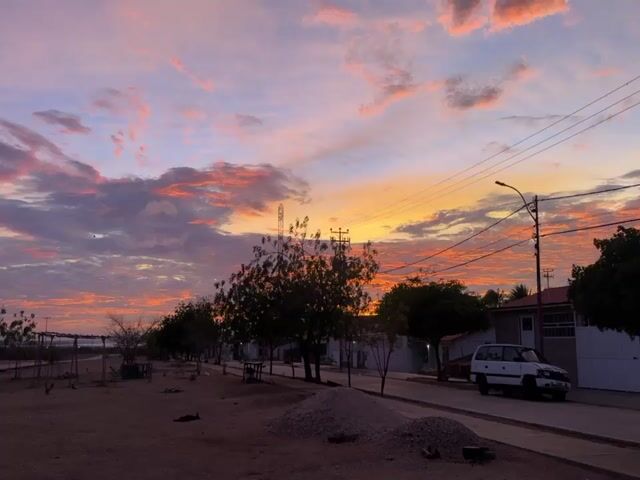 The video captures a sunset scene in a seemingly rural or suburban area. The sky is painted with vibrant colors of orange, pink, and purple clouds, indicating the sun is setting or has just set. In the foreground, there is a dirt road or path with some scattered debris, including piles of gravel. Several trees with dark foliage are planted along the side of the road, casting shadows. Beyond the trees, there are houses or buildings with white walls and reddish roofs. A tall communication tower or structure is visible in the distance. Electrical poles and wires stretch across the sky. On the right side of the road, a white vehicle, possibly a van or SUV, is parked. The overall atmosphere suggests a quiet evening in a developing area. The video is composed of still shots, likely from a camera or phone, with slight camera movements between each frame. There are no people or animals visible in the frame.