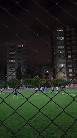 The video shows an artificial turf soccer field at night. A chain-link fence is in the foreground, slightly blurring the view of the field and players. Several individuals, dressed in athletic attire, are playing soccer. Two tall apartment buildings are visible in the background, with some windows illuminated. The sky is dark, suggesting it is nighttime.