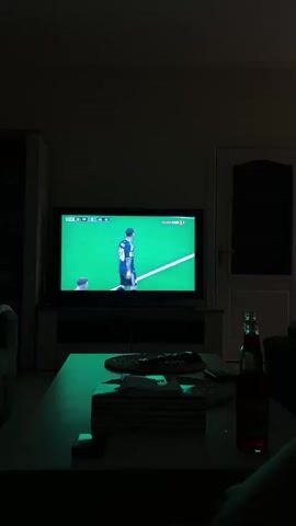 The video shows a living room scene in low light. A television is on, displaying a soccer match. On the field, a player wearing jersey number 10 is visible. In the foreground, a coffee table holds a box of tissues, a remote control, and a bottle with a red liquid. A person's leg is visible in the bottom right corner, suggesting someone is seated on a couch. The room has a door on the right side and a tall cabinet on the left. The lighting is dim, with a green or blue hue cast by the television screen.