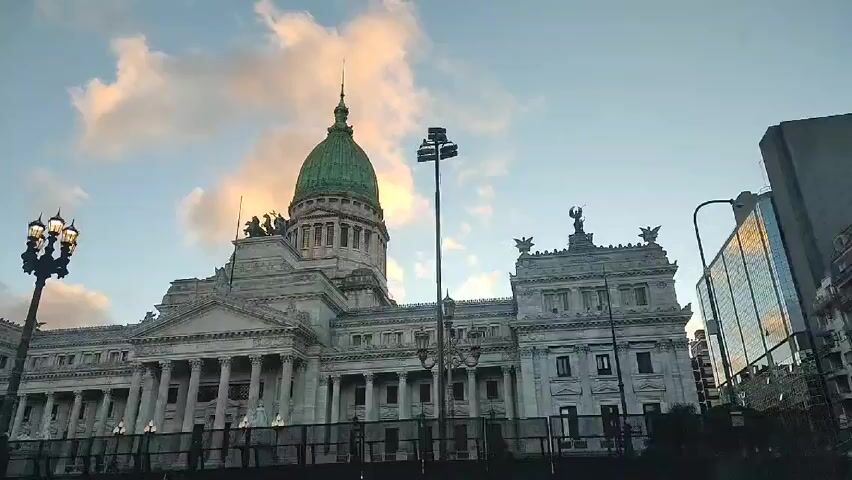 The video shows a daytime shot of the Palacio del Congreso Nacional (Palace of the National Congress) in Buenos Aires, Argentina. The building is large and ornate, with a green dome and numerous columns. There are street lights and a modern building visible in the background, and the sky is partly cloudy.  The camera is static, offering a view of the building from across a street, possibly behind a fence. The audio contains voices and music.