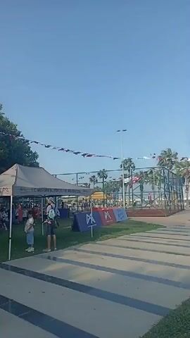 The video shows a wide outdoor shot on a sunny day. In the foreground, there are patterned concrete pathways and green grass. In the midground, a white tent is set up, with people gathered around and under it. Some people are children and appear to be participating in an activity. A flag is visible on a pole to the left of the tent, adorned with flags. Further back, trees line the area, and there are some parked vehicles. In the background, palm trees and a fence suggest the presence of a sports field or court. The scene appears to be a lively outdoor event.