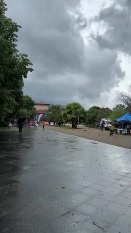 The scene is a wet, overcast day in a park. A paved path is visible in the foreground, reflecting the dull light from the gray sky. To the left, a person with an umbrella walks away from the camera. In the middle ground, figures are spaced along the path, some running, some walking, possibly participating in an event marked by orange cones. An ambulance and a blue tent are present on the right side of the path. Trees surround the area, and a building with banners is visible further in the background. The general impression is of a subdued outdoor event in inclement weather.